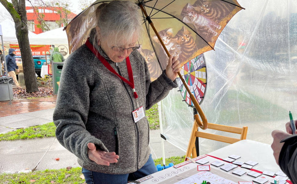 Woman playing a board game, wearing fleece and holding an umbrella, with transparent plastic sheeting forming the side of a booth, and rainy market stalls in the background.