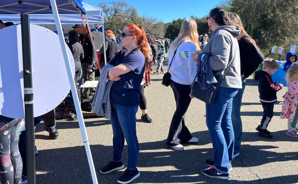 People and children standing in line at a festival booth with a prize wheel at the front.